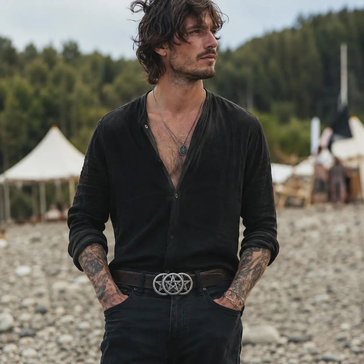 Man wearing a black shirt, pentagram belt buckle and black pants with tattoos, standing on a pebbly beach with tents and trees in the background.
