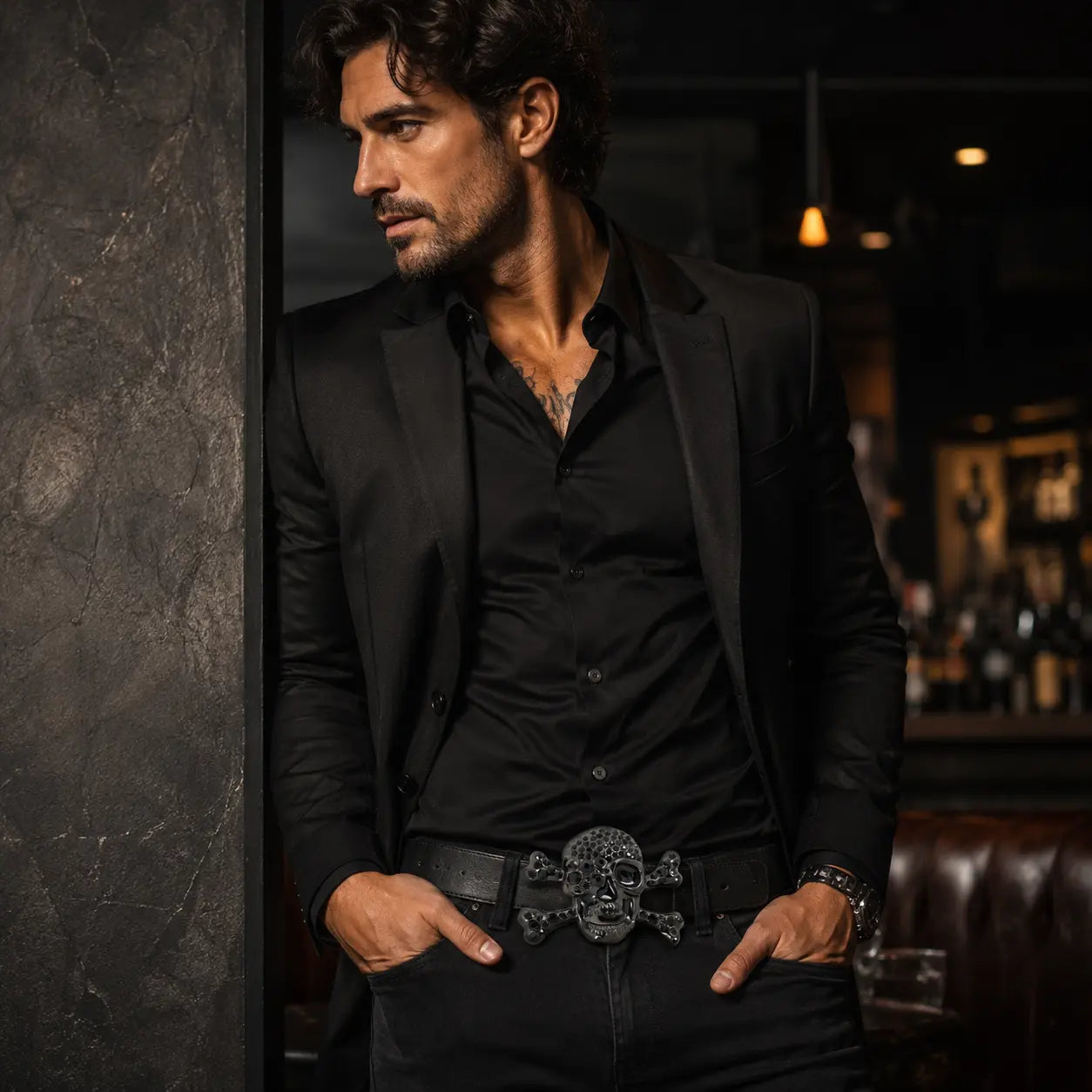 Man in a black suit with a skull belt buckle in a dimly lit room.