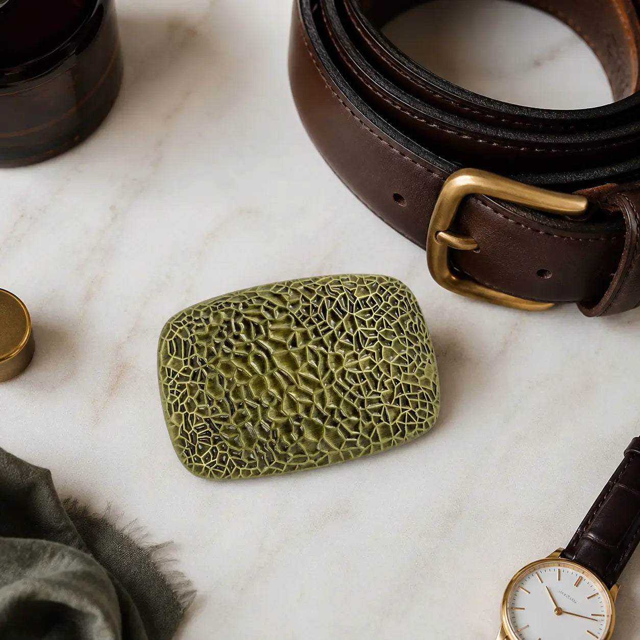 An olive brass coloured textured belt buckle with a rectangular shape and rounded corners, displayed on a white marble surface next to a brown leather belt, a watch, and other lifestyle accessories.