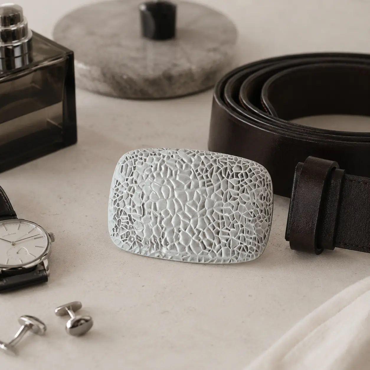 A textured, rectangular light gray belt buckle with a cracked or mosaic pattern, displayed on a light beige surface next to a dark brown leather belt, a perfume bottle, a watch, and silver cufflinks.