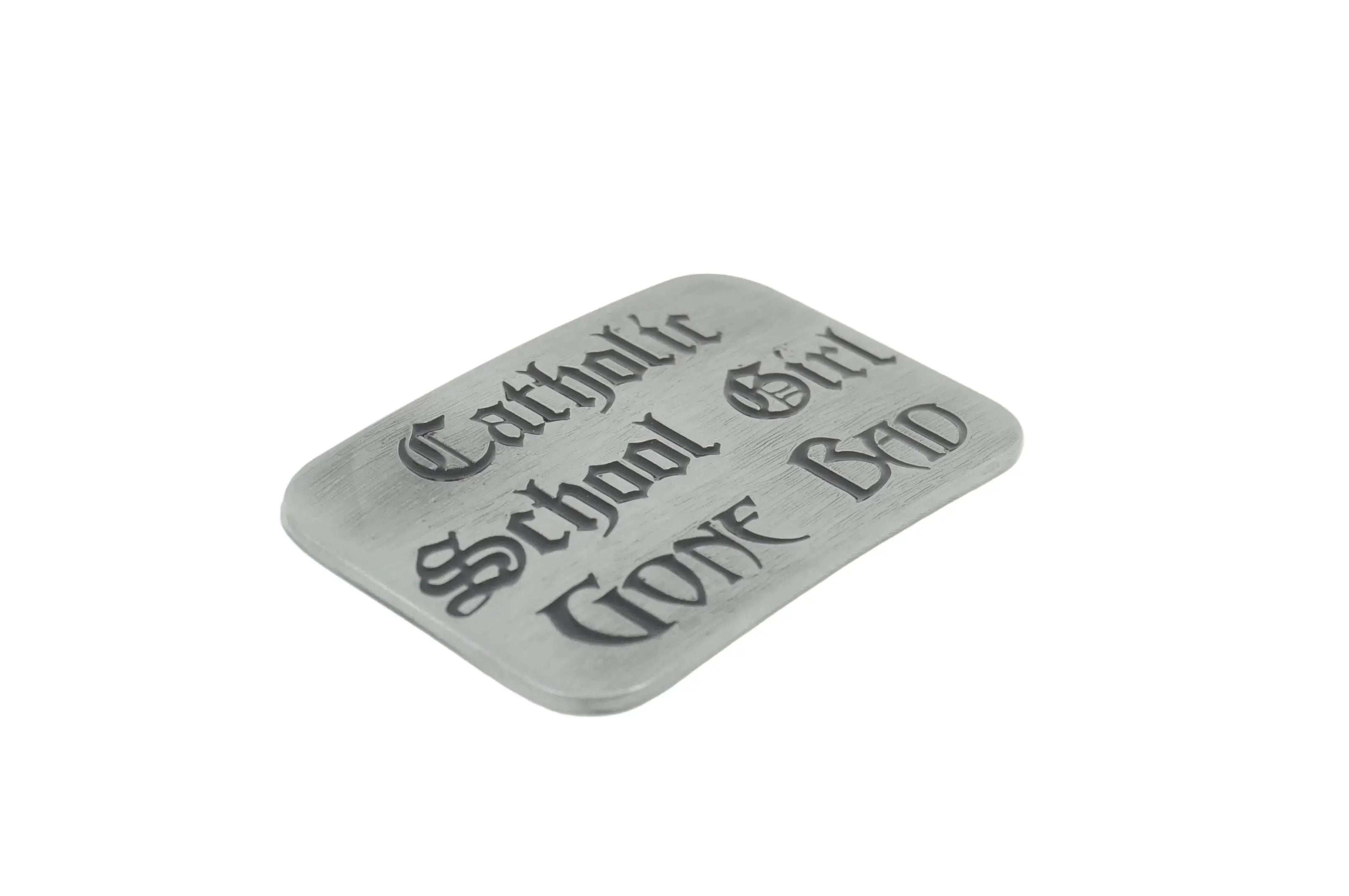 A rectangular gray metal belt buckle with rounded corners, featuring the gothic-style text 'Catholic School Girl WENT BAD' engraved on its front, set against a plain white background.