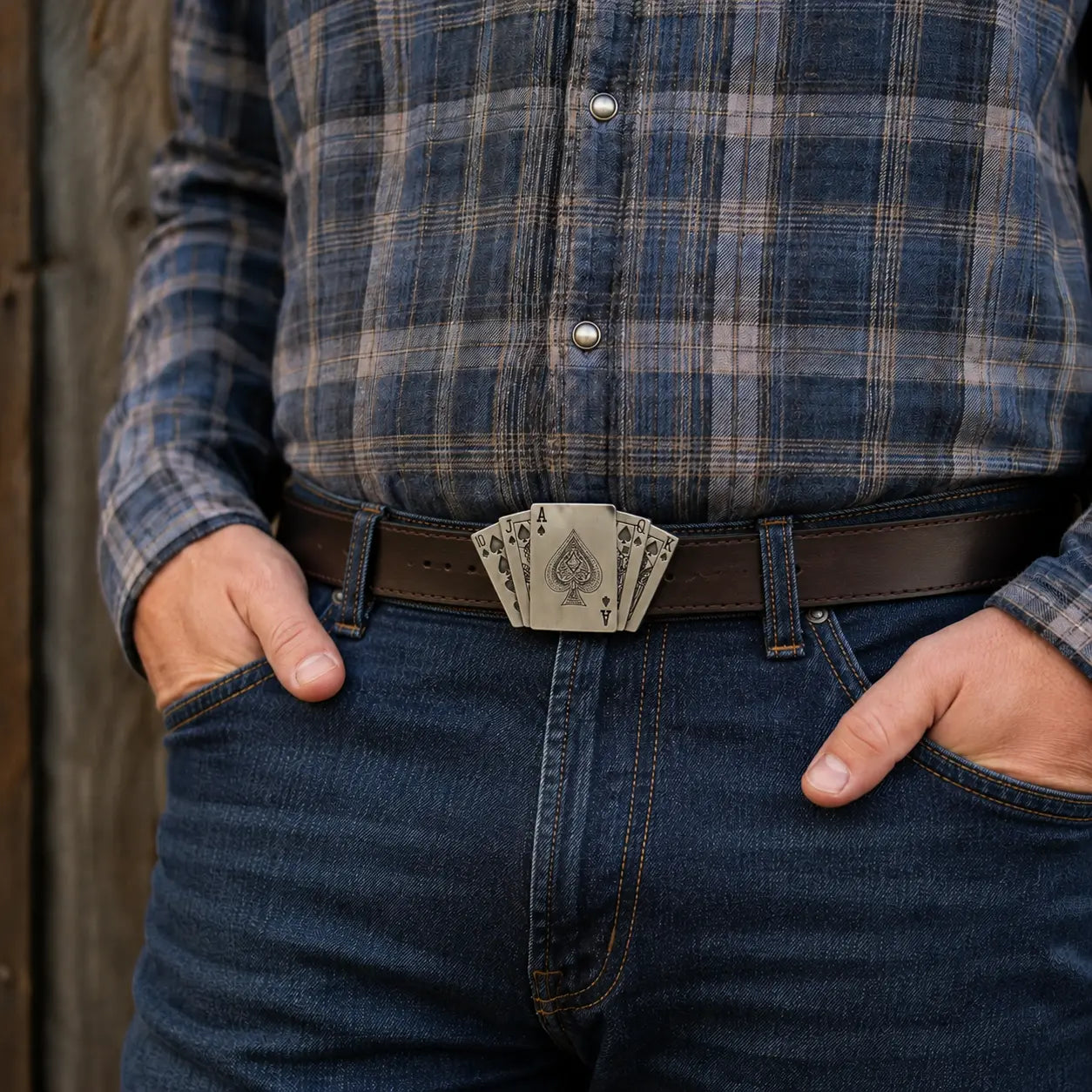 A close-up of a person wearing a blue and gray plaid flannel shirt, dark blue jeans, and a brown leather belt with a PLAYING CARDS metal buckle shaped like a fanned deck of playing cards.