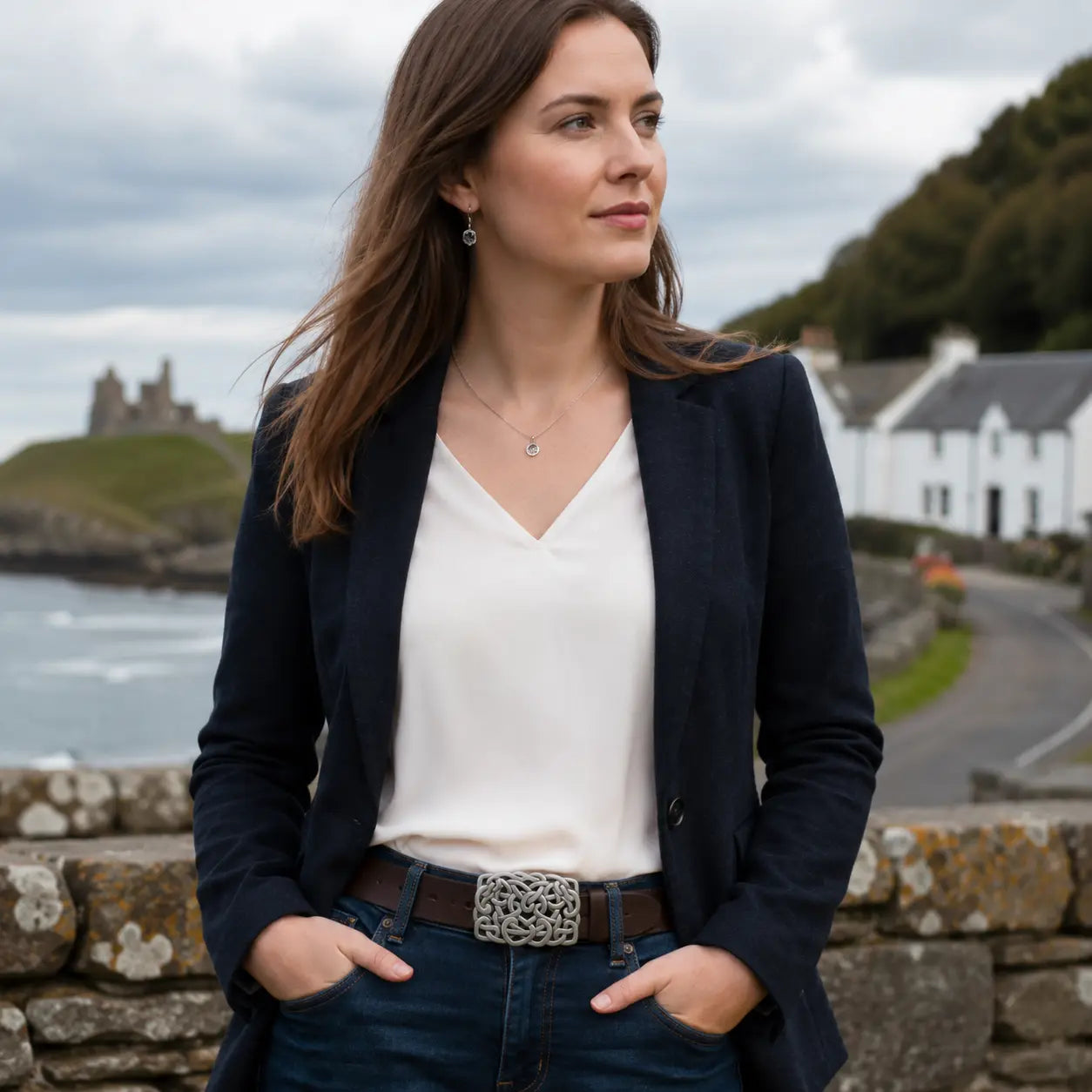 A woman wearing a dark navy blazer, white V-neck top, and dark blue jeans with a silver belt buckle stands outdoors near a stone wall, with a coastal village and castle in the background.