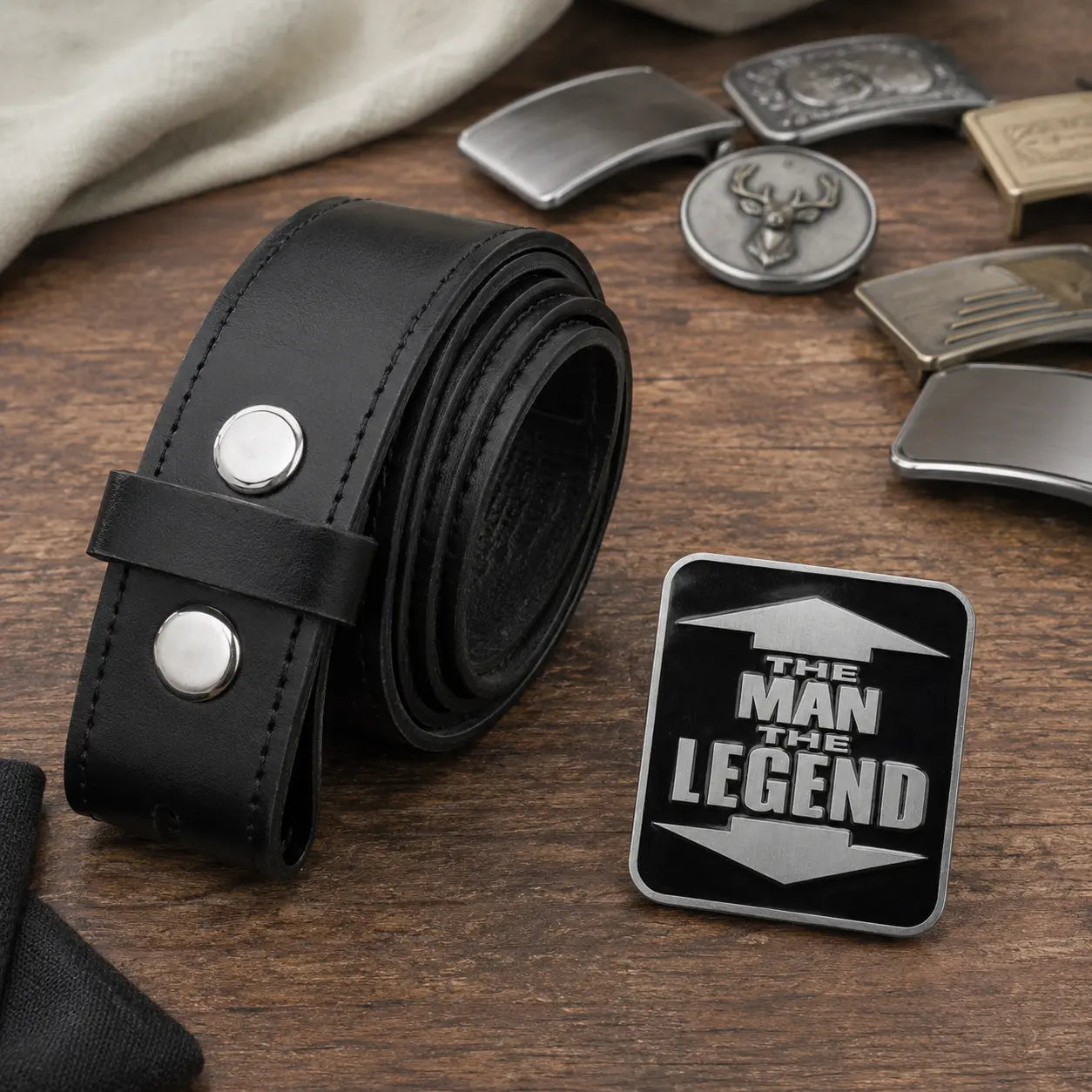 A black leather belt with silver snap buttons is displayed on a wooden surface next to a black belt buckle engraved with the text 'THE MAN THE LEGEND'. Several other metallic belt buckles are scattered in the background.