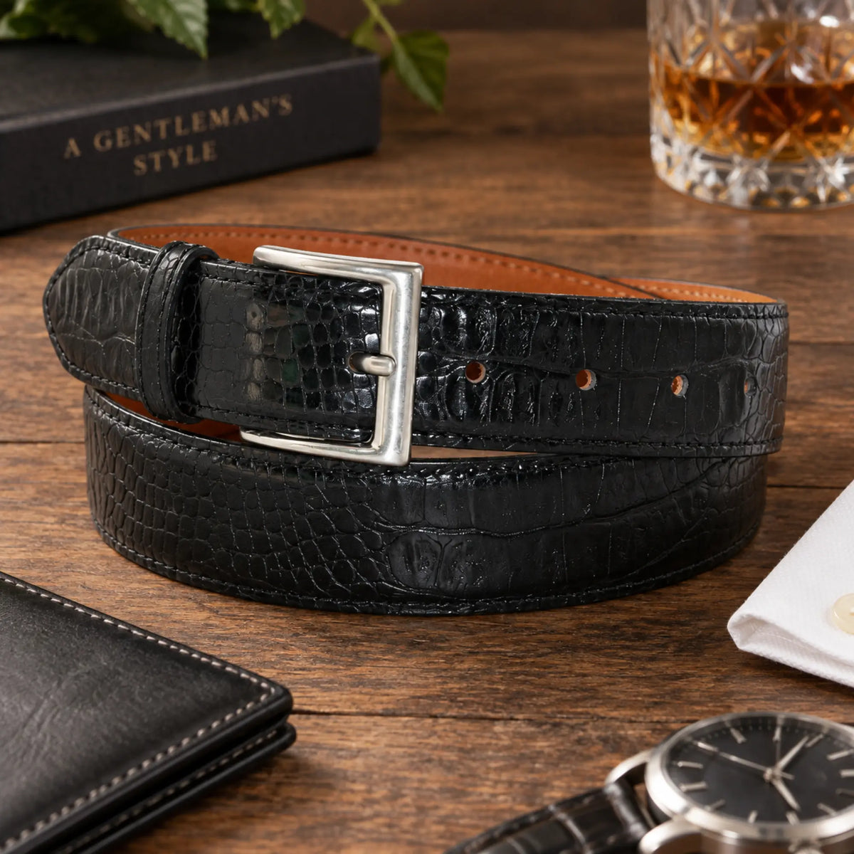 A black crocodile-embossed leather belt with a silver buckle, coiled on a wooden tabletop with lifestyle props including a watch, wallet, book, and a glass of whiskey.