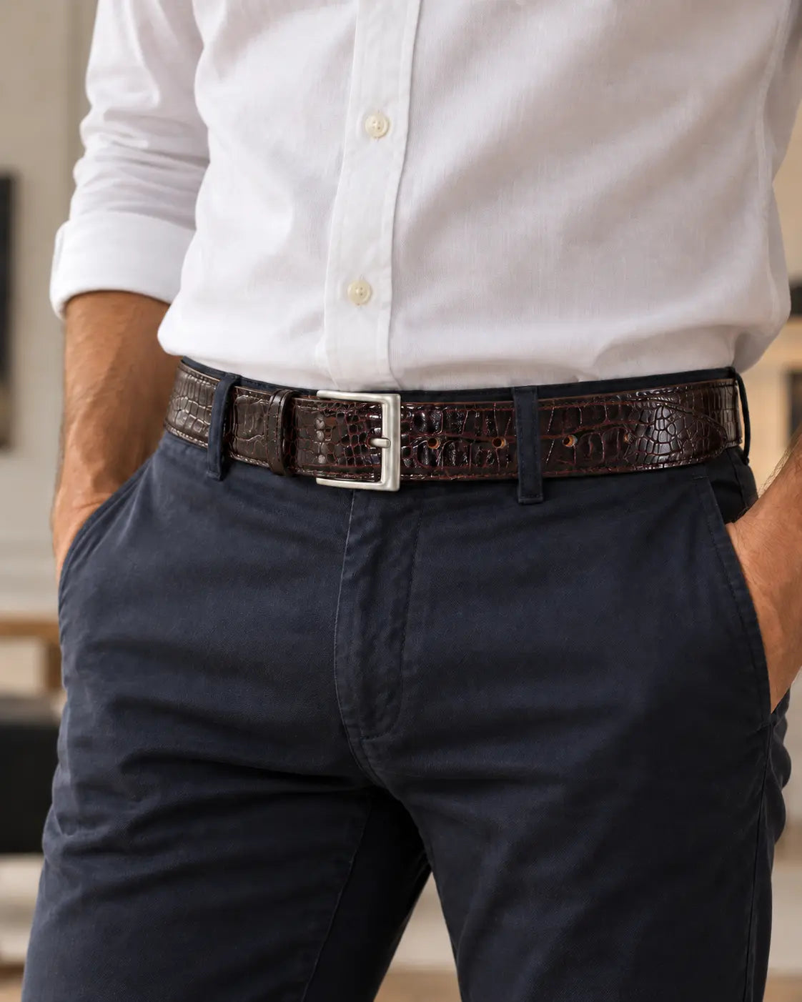 A man wearing a dark brown crocodile-embossed belt with a silver buckle, paired with a white button-down shirt and navy blue chinos.