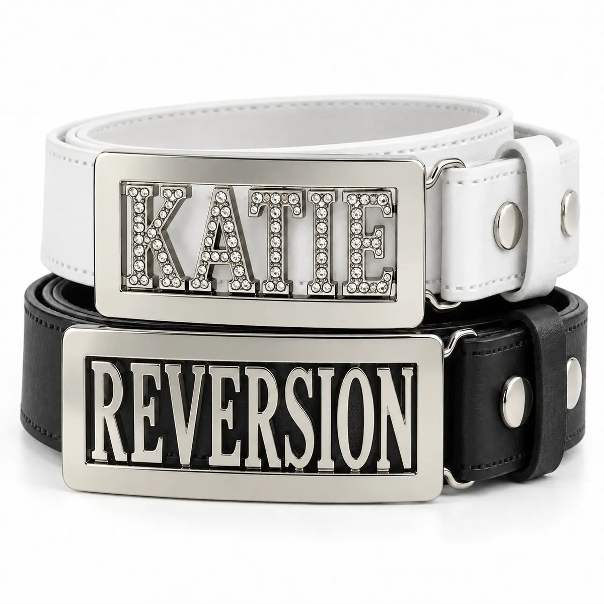 Two leather personalised belts stacked on a white background: a white belt with a silver rectangular buckle featuring the name 'KATIE' in rhinestone letters, and a black belt with a matching silver rectangular buckle featuring the name 'REVERSION'.