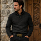 Man wearing a dark grey sweater with a bronze celtic belt buckle in front of a wooden door and stone wall in the background