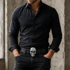 Man wearing a black shirt and dark jeans with a skull belt buckle against a neutral background