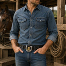 Man wearing a blue denim shirt and jeans wearing a horseshoe belt buckle standing in front of a horse in a barn.