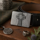 Silver Celtic cross belt buckle with matching pendant and ring on a wooden surface.
