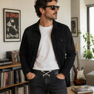 A man wearing a black denim jacket over a white t-shirt and dark jeans with a guitar belt buckle, standing in a room with framed Led Zeppelin posters, a record shelf, and a guitar in the background.