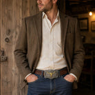 Man wearing a brown blazer, white shirt, and blue jeans with a large belt buckle in a rustic setting.