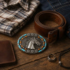 A western-style belt buckle featuring a raised horse head motif with turquoise and brown accents, displayed on a rustic wooden surface alongside a brown leather belt, folded plaid shirt, denim fabric, a wallet, rings, and a cord accessory.