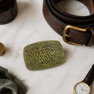 An olive brass coloured textured belt buckle with a rectangular shape and rounded corners, displayed on a white marble surface next to a brown leather belt, a watch, and other lifestyle accessories.