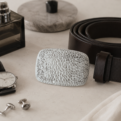 A textured, rectangular light gray belt buckle with a cracked or mosaic pattern, displayed on a light beige surface next to a dark brown leather belt, a perfume bottle, a watch, and silver cufflinks.