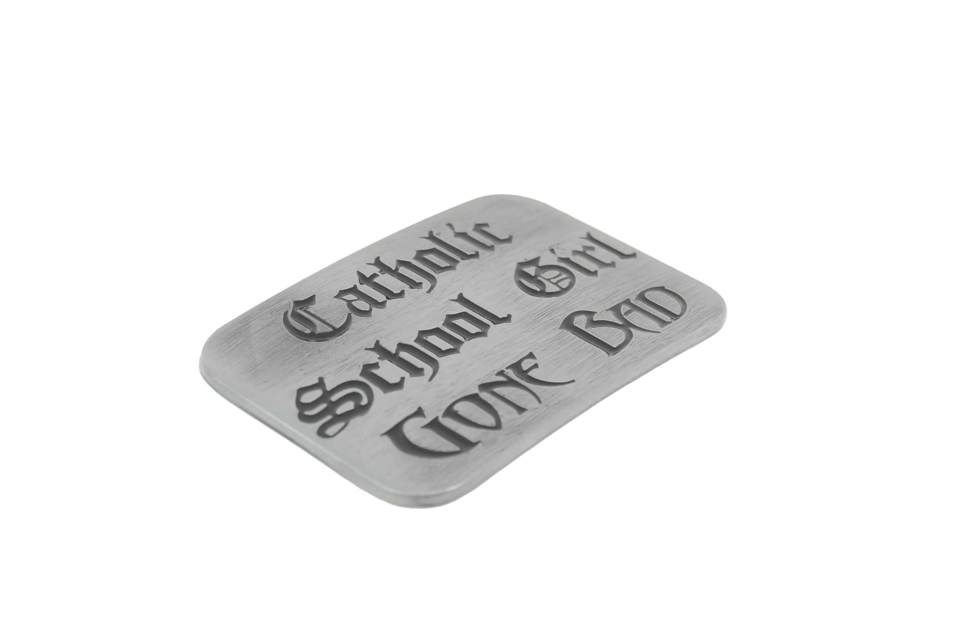 A rectangular gray metal belt buckle with rounded corners, featuring the gothic-style text 'Catholic School Girl WENT BAD' engraved on its front, set against a plain white background.