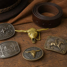 A styled flat-lay of western belt buckles and a brown leather belt on a wooden surface with draped brown fabric. The belt features a rolled dark brown leather loop with silver rivets and a stitched edge.