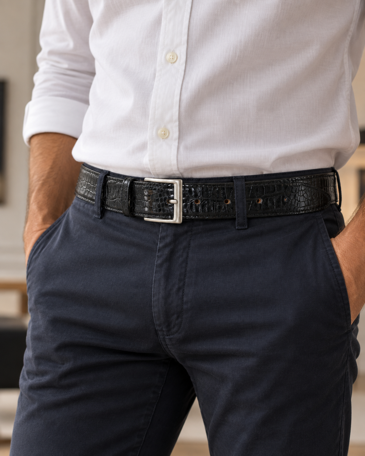 A man wearing a black crocodile-embossed belt with a silver buckle, paired with a white button-down shirt and navy blue trousers.