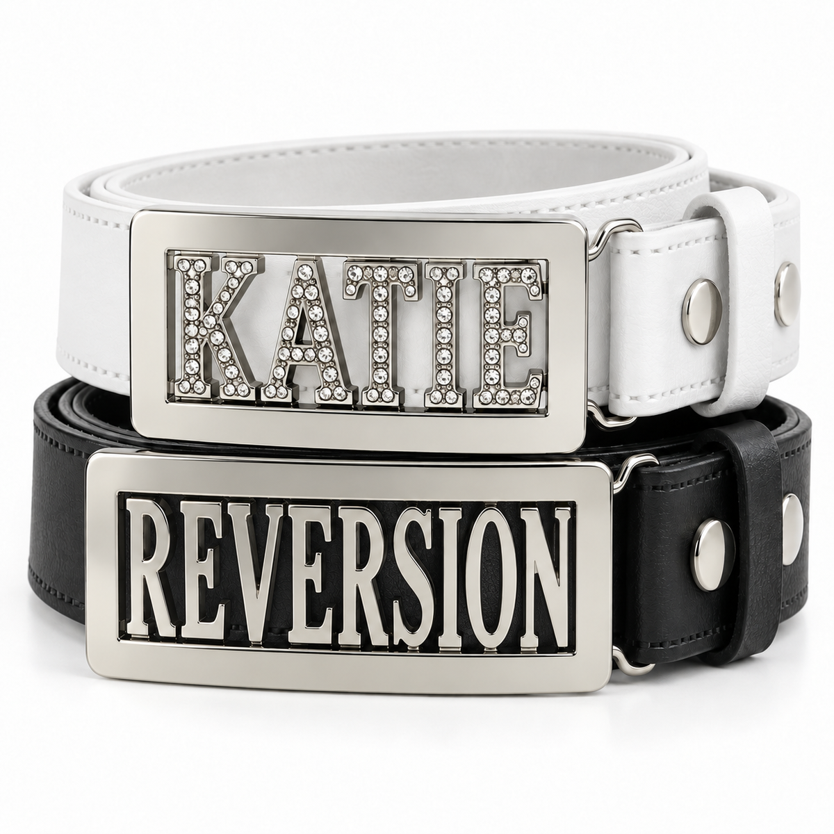 Two leather personalised belts stacked on a white background: a white belt with a silver rectangular buckle featuring the name 'KATIE' in rhinestone letters, and a black belt with a matching silver rectangular buckle featuring the name 'REVERSION'.
